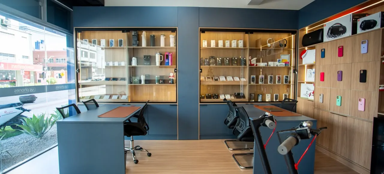 Interior da VS Phones: vitrines de madeira e azul-marinho com iPhones, AirPods, capas, caixas JBL e copos Stanley, e a rua do Centro de Criciúma pela vitrine